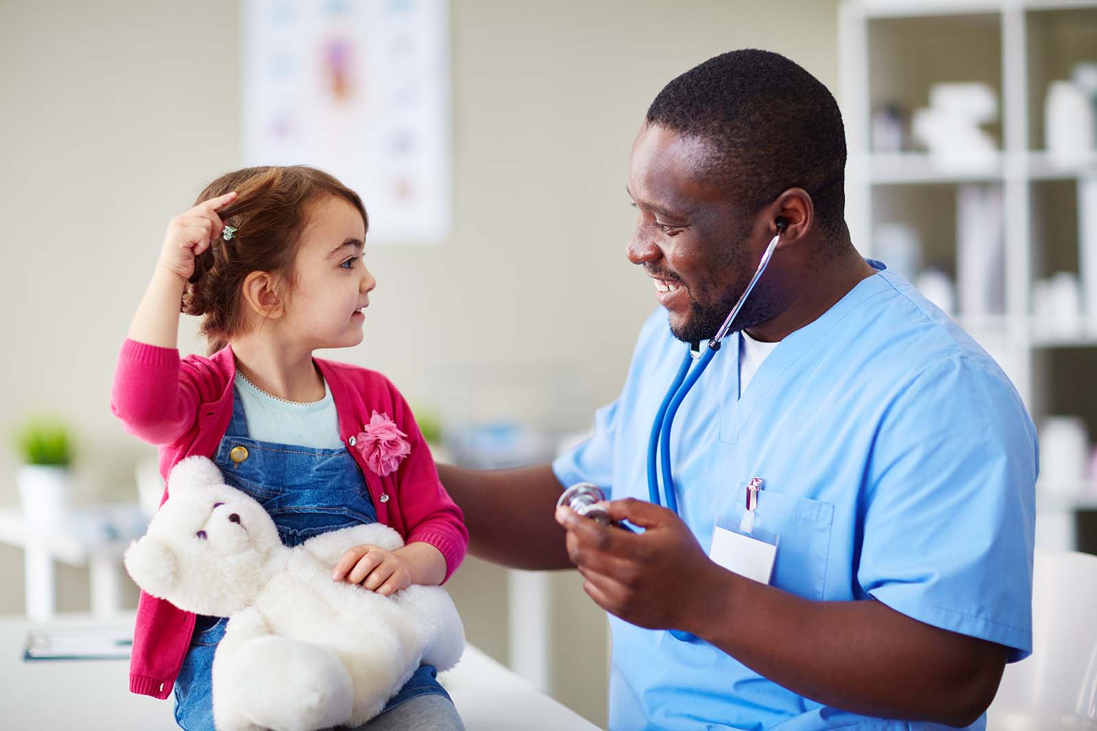 A smiling male nurse talking to a young child holding a large stuffed bear (the child is holding the bear, not the nurse)