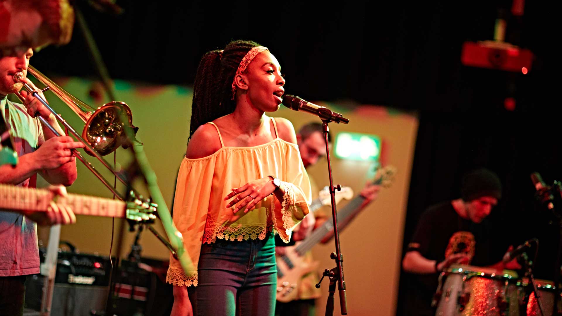 A female student sings into a microphone while other students play instruments around her.