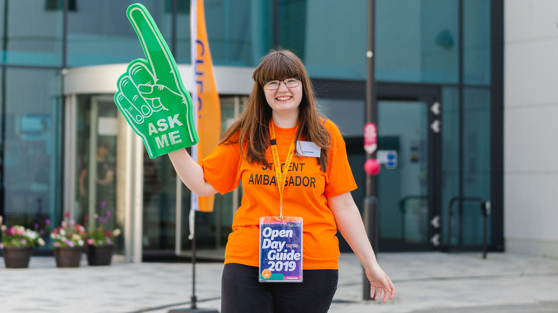 A student ambassador outside Oastler on Open Day with a green ask me foam hand