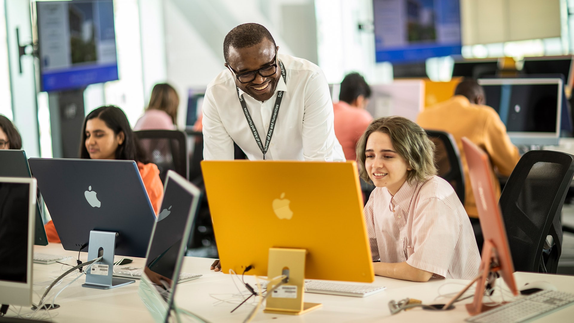 An smiling academic stood next to a student working on a desktop computer. The academic is looking down at the student's work.