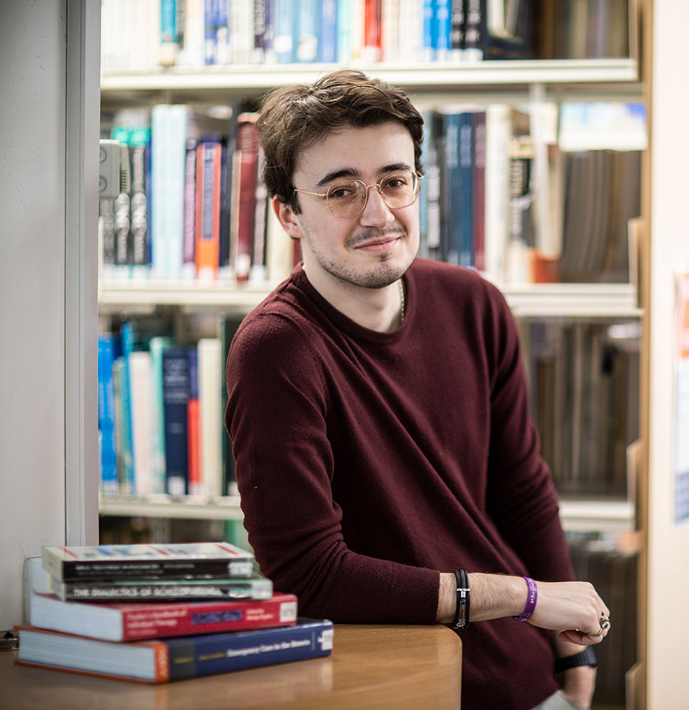 PhD student Tom Williams, leaning against a desk with some books on it, in the library.