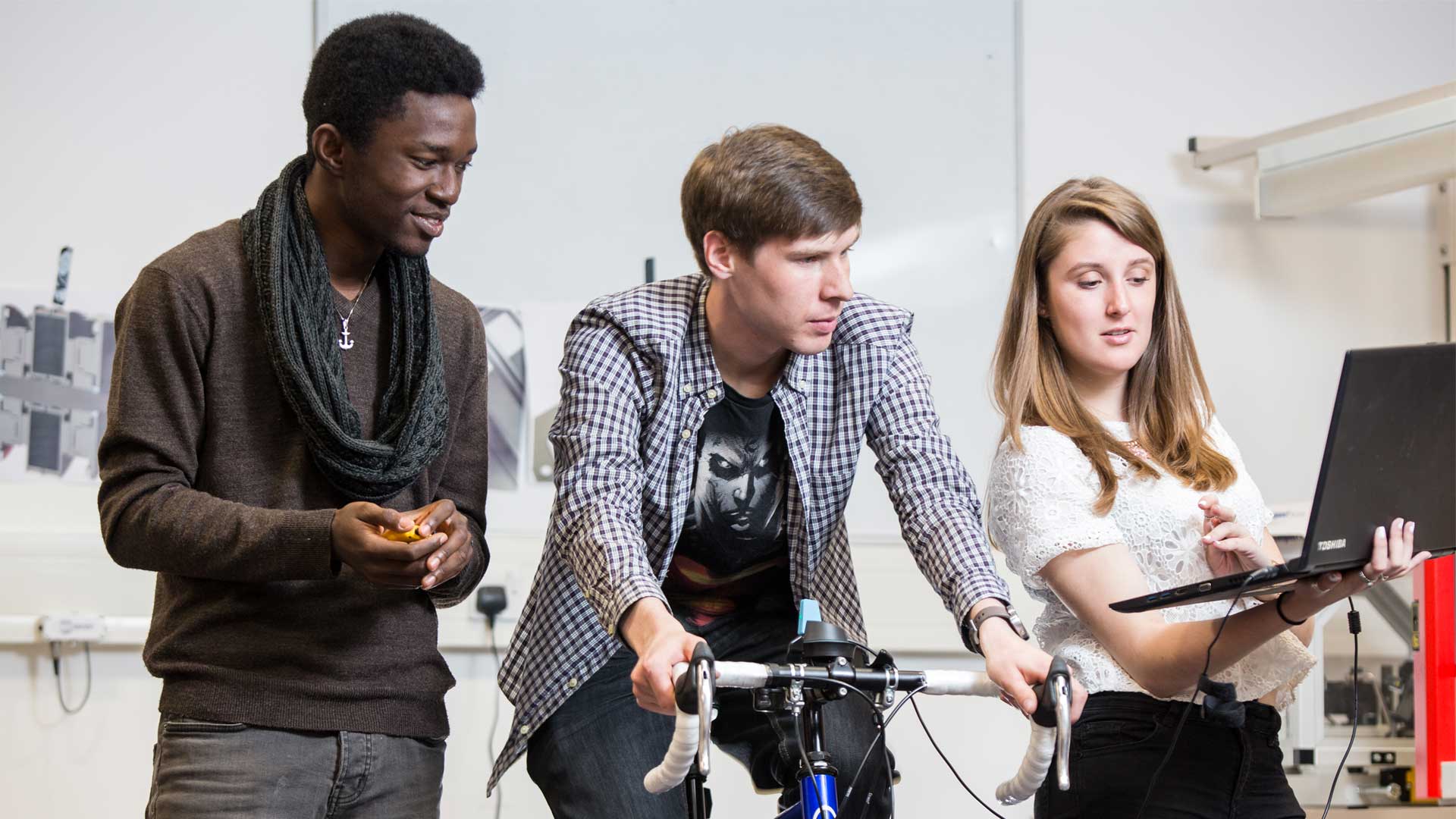 2 students stand around a bike while running a test. Another student is sat on the bike.